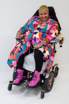 A woman with short, buzzed hair wearing a colorful shawl and pink boots smiles while seated in a power wheelchair