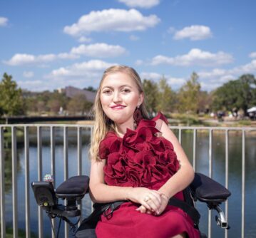 A young woman with blonde hair wearing a red dress sits in a power wheelchair in front of a lake