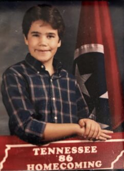 A young man in a plaid shirt smiles in front of a Tennessee flag and a sign that says Tennessee 86 Homecoming