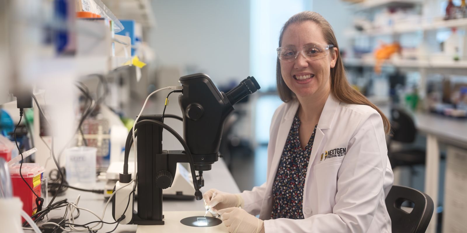 A young female scientist in a lab coat sits next to a high powered microscope in a research lab