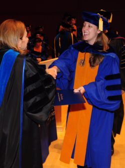 A young woman in graduation gown and scarf and hat walks across the stage to accept her PhD diploma