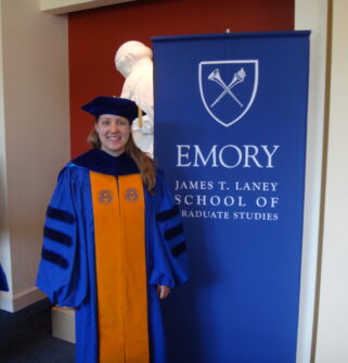 A young woman in graduation gown with scarf and hat stands in front of an Emory sign