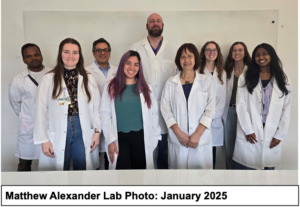 Matthew Alexander, PhD, stands in the back row of a coed group of nine scientists in white lab coats. He is a head taller than the other scientists and has a bald head and a full dark brown beard.