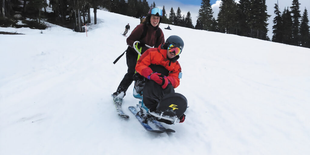 Sean Marihugh smiles as he slides down a ski slope in a sit-ski with a partner on skis holding onto the back of his sit-ski.