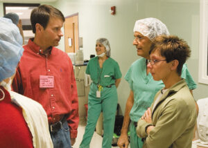 A group of people stands in a hospital corridor. Most are wearing scrubs and head covers. One man and one woman are wearing plain clothes, and the man has a name tag that says “Parent.”