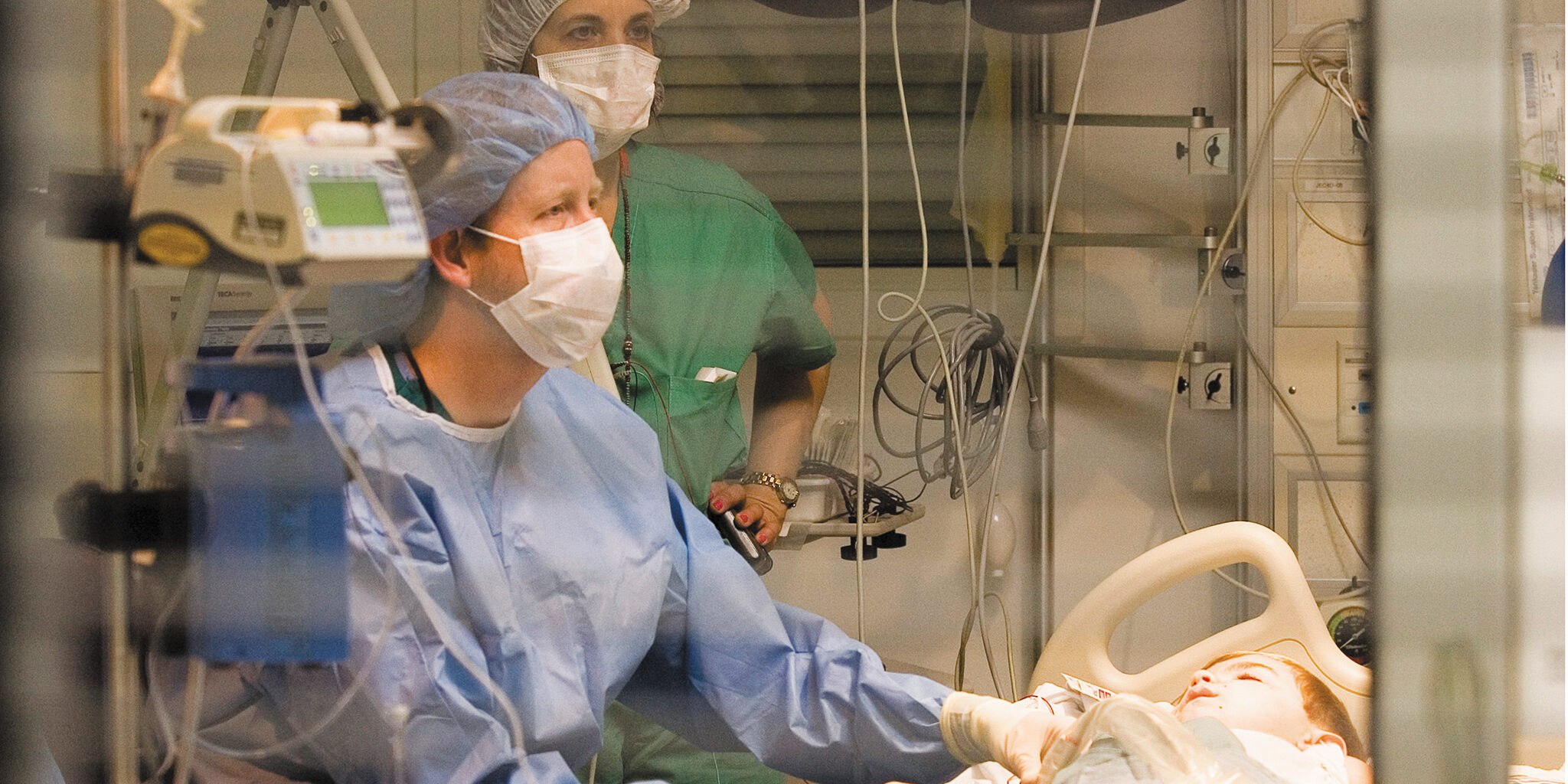 Inside a hospital room, two healthcare providers wearing scrubs, gowns, masks, and head covers stand beside a bed where a young boy is lying down.