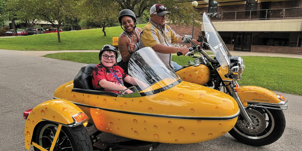 An MDA volunteer gives campers rides on a motorcycle with a sidecar painted to look like a wedge of cheddar cheese at MDA Summer Camp.