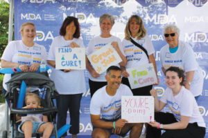 A group of seven adults and one toddler in a stroller are gathered outdoors wearing matching T-shirts that say “Muscle Walk.” Some hold signs with encouraging sayings, like “Great Job” and “Find a Cure.”