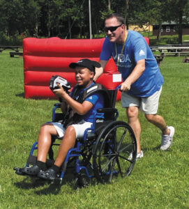 A man in a blue MDA T-shirt pushes a boy in a wheelchair through a grassy field on a sunny day.