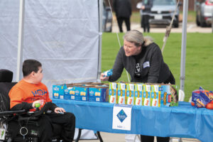 At an outdoor table set up with boxes of granola bars and Rice Krispies Treats, a woman offers a snack to a boy in a power wheelchair.