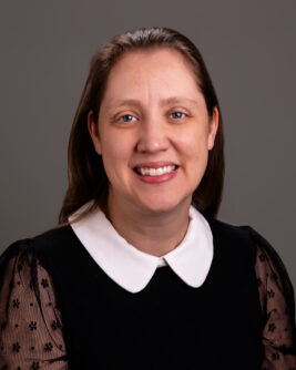 A woman with brown hair and red lipstick wearing a black dress shirt with a white collar smiles against a gray backdrop