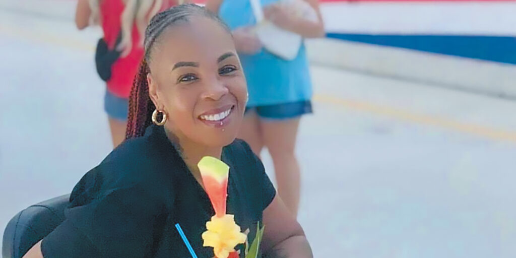 Mashauna Black, a woman with brown skin and black and red braids pulled back into a ponytail, sits in scooter holding a tropical drink.