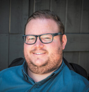 Headshot of Tyler Dukes, a man with short reddish-brown hair and beard, light freckled skin, and black glasses.