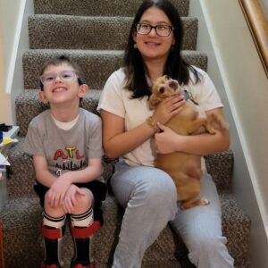 A young boy wearing leg braces and a teenage girl sit on carpeted stairs holding a small brown dog.