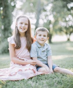 A girl in a pink summer dress sits in the grass with her arm around a younger boy wearing a blue Polo shirt and shorts.