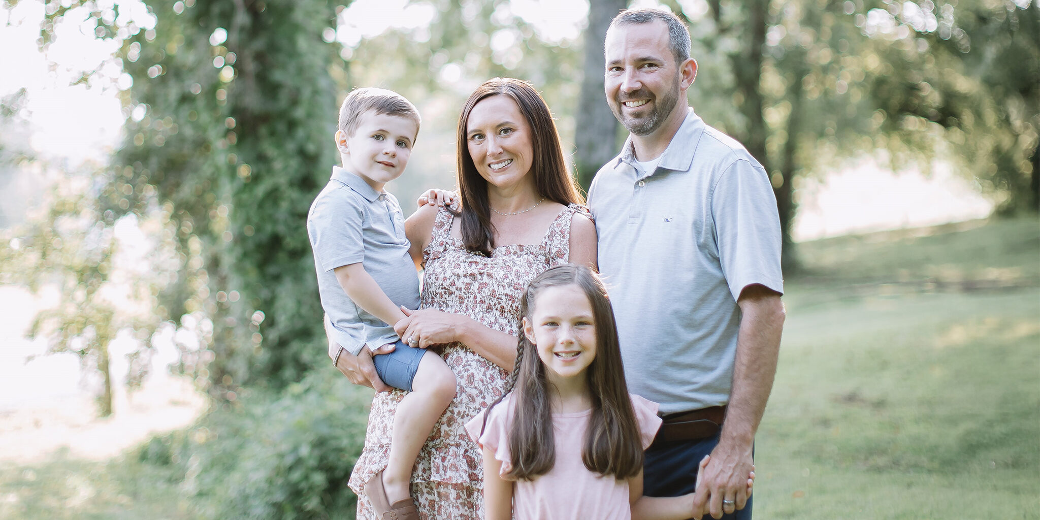 A family of four, consisting of a mother holding a toddler boy and a father holding his daughter’s hand, poses in an outdoor setting.