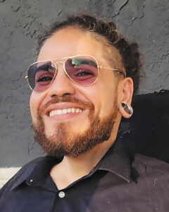 Closeup of Caeser Chacon-Carro, a man with short, curly brown hair and beard and light skin, wearing gold aviator sunglasses and silver ear gauges.