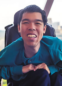 Closeup of Justin Moy, an Asian man with short black hair and light skin, sitting in a black power wheelchair.