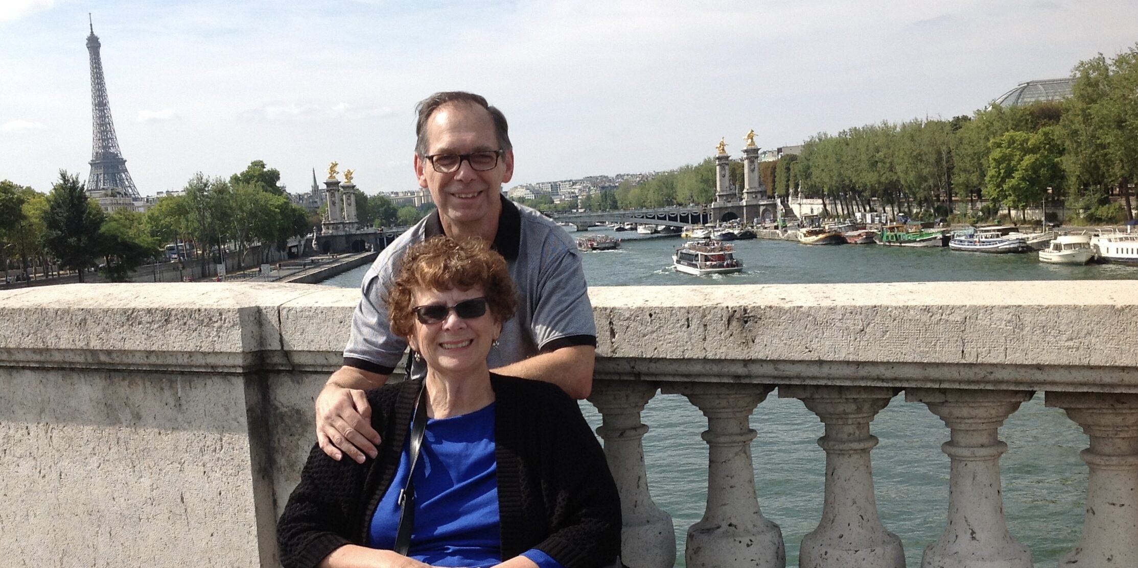 Barbara and Jim Twardowski pose on a bridge over the Seine in Paris with the Eiffel Tower visible in the background. Barbara is sitting in a wheelchair and Jim is standing behind her.