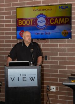 A man in a black polo stands in front of a podium with a sign that reads Boot Camp MDA behind him