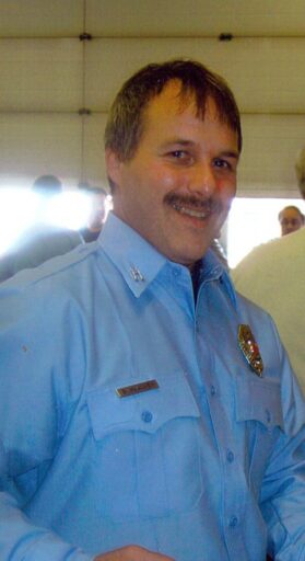 A fire fighter in a blue button down with brown hair and a mustache smiles at the camera