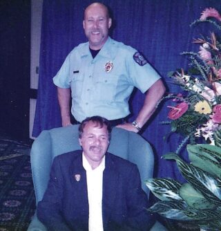 A man in a fire fighter shirt poses behind a man seated in a green chair