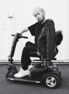 Black-and-white shot of Danny Kurtzman sitting in a mobility scooter with his head cocked and shoulders turned toward the camera, a black leather motorcycle draped over one shoulder.