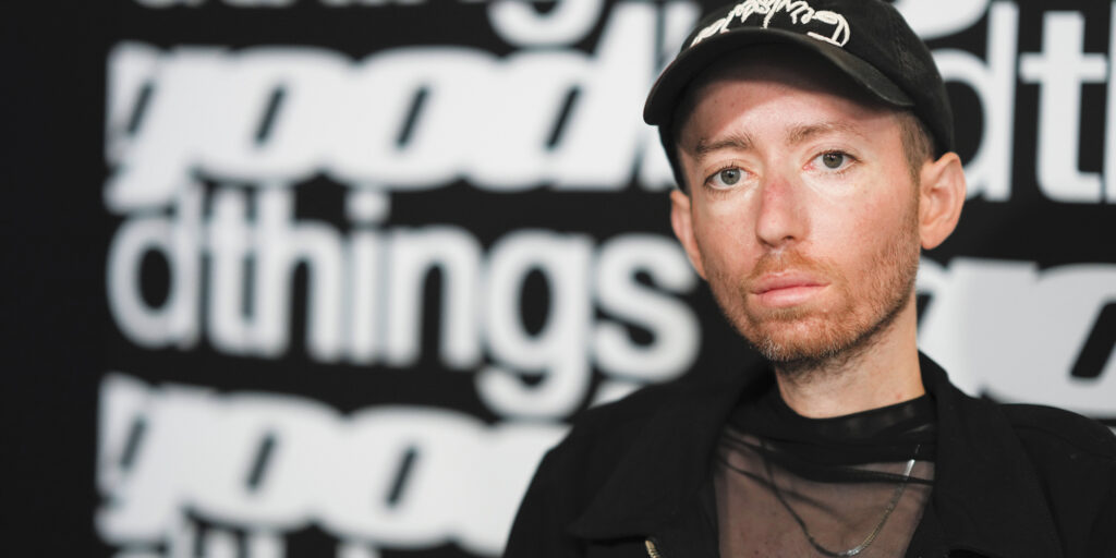 Close up of actor Danny Kurtzman at a screening of the movie Good Bad Things. He is looking straight at the camera, wearing a black cap and black shirt and jacket in front of a backdrop printed with the movie title.