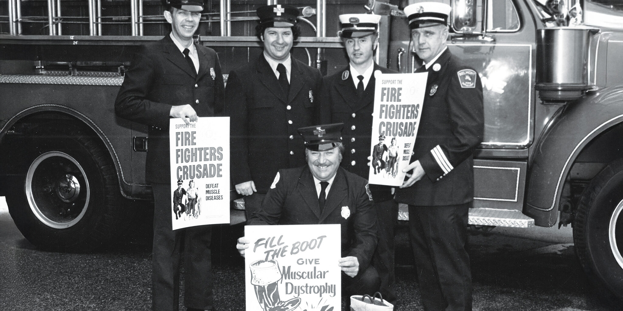 1950s black-and-white photo of five fire fighters in uniform holding signs saying “Fire Fighters Crusade” and “Give Muscular Dystrophy a Kick” in front of an open-cab ladder truck.