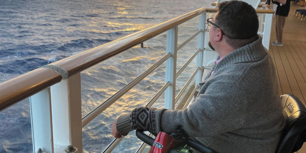 Matt Curcio sitting in a mobility scooter on the deck of a cruise ship looking over the water at the sunset.