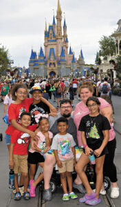 Matt Curcio sits in a mobility scooter, with his wife standing on his left, in the middle of a group of six kids who appear to range in age from 6 to teens, with Disney Worlds’s Cinderella Castle behind them.