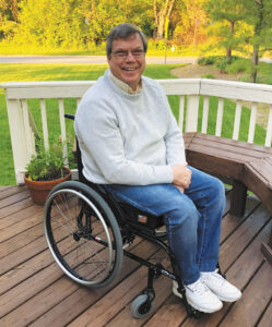 David Capozzi smiles at the camera while sitting in a manual wheelchair on an outdoor deck on a sunny day.