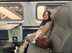 Leah Zelaya wearing bilateral leg braces in accessible seating on an Amtrak train. In front of her there is legroom and a foldaway table with the International Symbol of Accessibility on it.