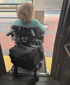View from behind of Mindy Henderson driving her power wheelchair down a short metal ramp to deboard the train.