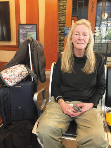 Vanessa O’Connell, dressed comfortably for travel, sitting in the waiting room at an Amtrak station with a suitcase, carryon bag, and jacket stacked next to her.
