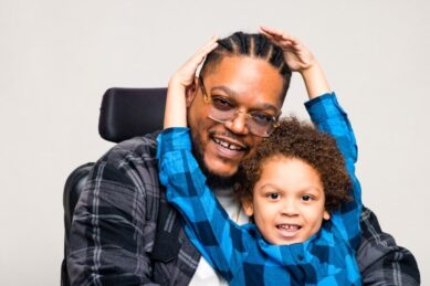 A man wearing glasses and sitting in a wheelchair holds his son in his lap