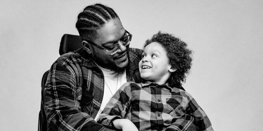 A dad in a wheelchair wearing glasses smiles down at the young child he is holding in his lap