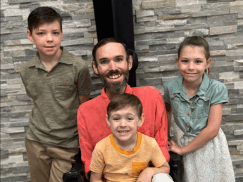 A man in a wheelchair smiles while his three children gather around him