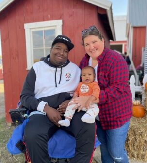 A man in a wheelchair and his wife standing beside him hold a young infant in front of a barn