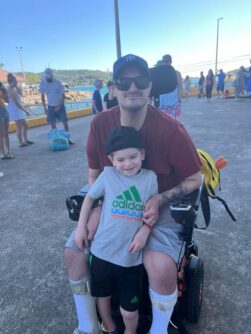 A young dad in a baseball cap and sunglasses smiles in a wheelchair with his young son leaning against him.