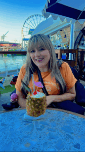Sory Rivera sitting at an outdoor table with a tropical drink in a pineapple in front of her and water and a Ferris wheel in the background.
