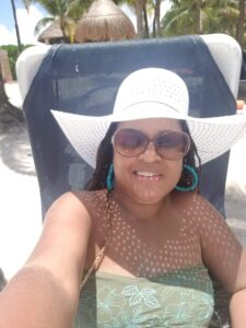 Selfie of Tamara Blackwell, wearing a wide-brimmed sun hat and sunglasses, reclining in a beach lounger with palm trees and a thatched hut in the background.