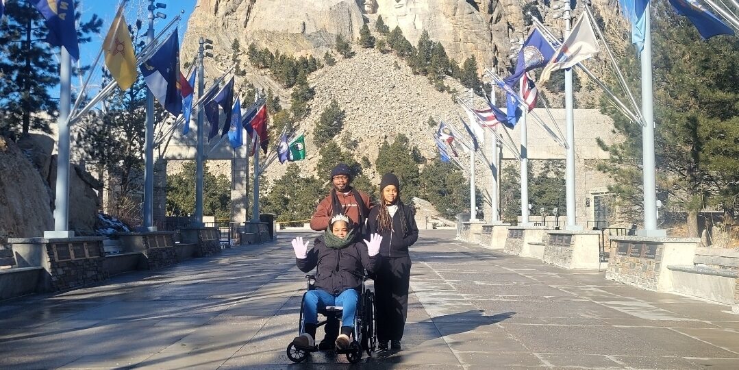 Tamara Blackwell, in a wheelchair, and her two travel companions pose with Mt. Rushmore in the background.