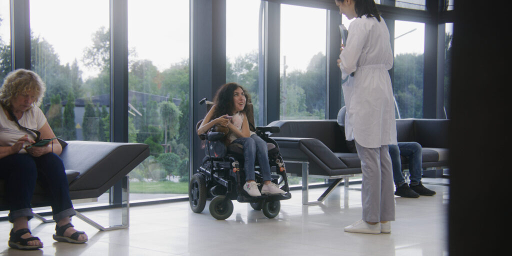 Woman in electric wheelchair waits for consultation with doctor in clinic waiting area. Nurse invites patient with disability on procedures. Medical staff and people in modern medical facility lobby.