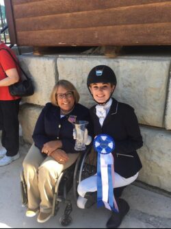 A young woman in equestrian gear and holding ribbons crouches next to a woman in a wheelchair
