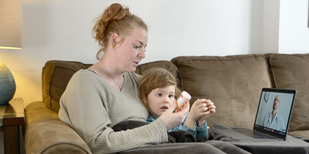 A mother is sitting on a couch with her sick daughter. They are they are having a video call on a laptop computer with a male doctor.