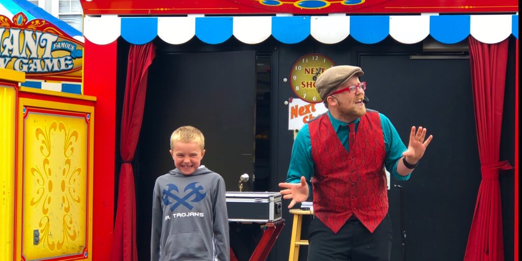 A magician in a red vest and hat stands on as stage with a young boy