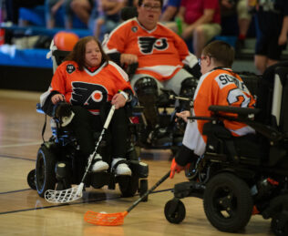 Three power hockey players in orange jerseys on the court. 