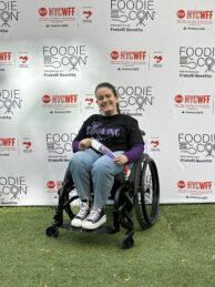 Gabby DiSalvo, seated in her wheelchair wearing a T-shirt that says Cooking on Wheels, poses holding a purple spatula in front of an event backdrop printed with logos for FoodieCon and New York City Wine and Food Festival.