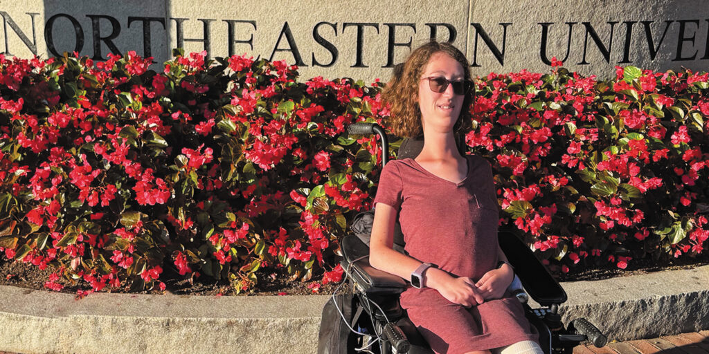 Graduate student Naomi Hess sitting in a power wheelchair in front of a Northeastern University sign in Boston.